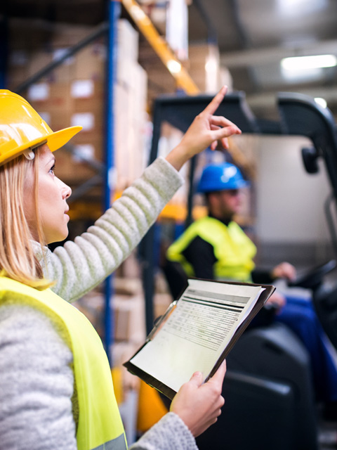 Warehouse worker in a yellow hard hat and vest points to something while holding a clipboard, with a forklift operator in the background.